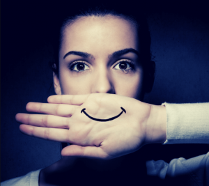 Na imagem: uma pessoa chorando com um papel na frente do rosto onde está desenhado um sorriso