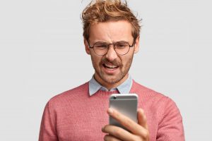 Na imagem: Homem ruivo com óculos redondo fazendo cara feia enquanto olha para uma conversa ruim no celular.