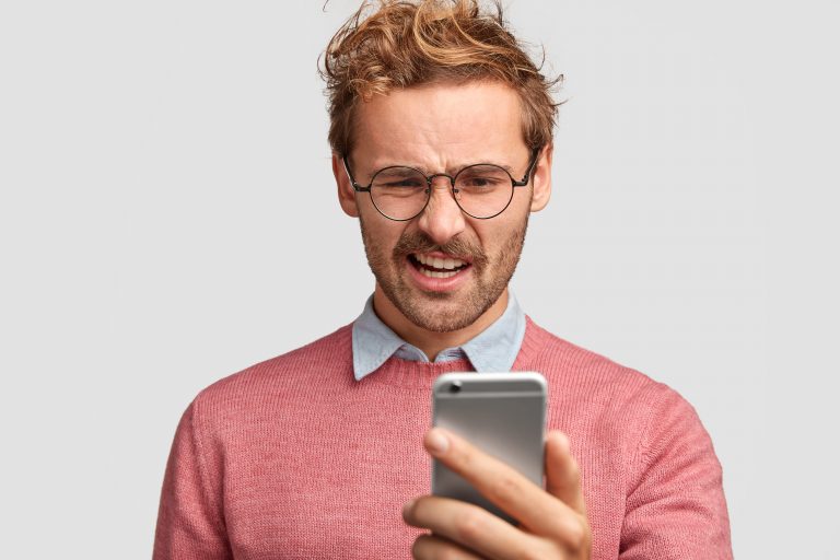 Na imagem: Homem ruivo com óculos redondo fazendo cara feia enquanto olha para uma conversa ruim no celular.